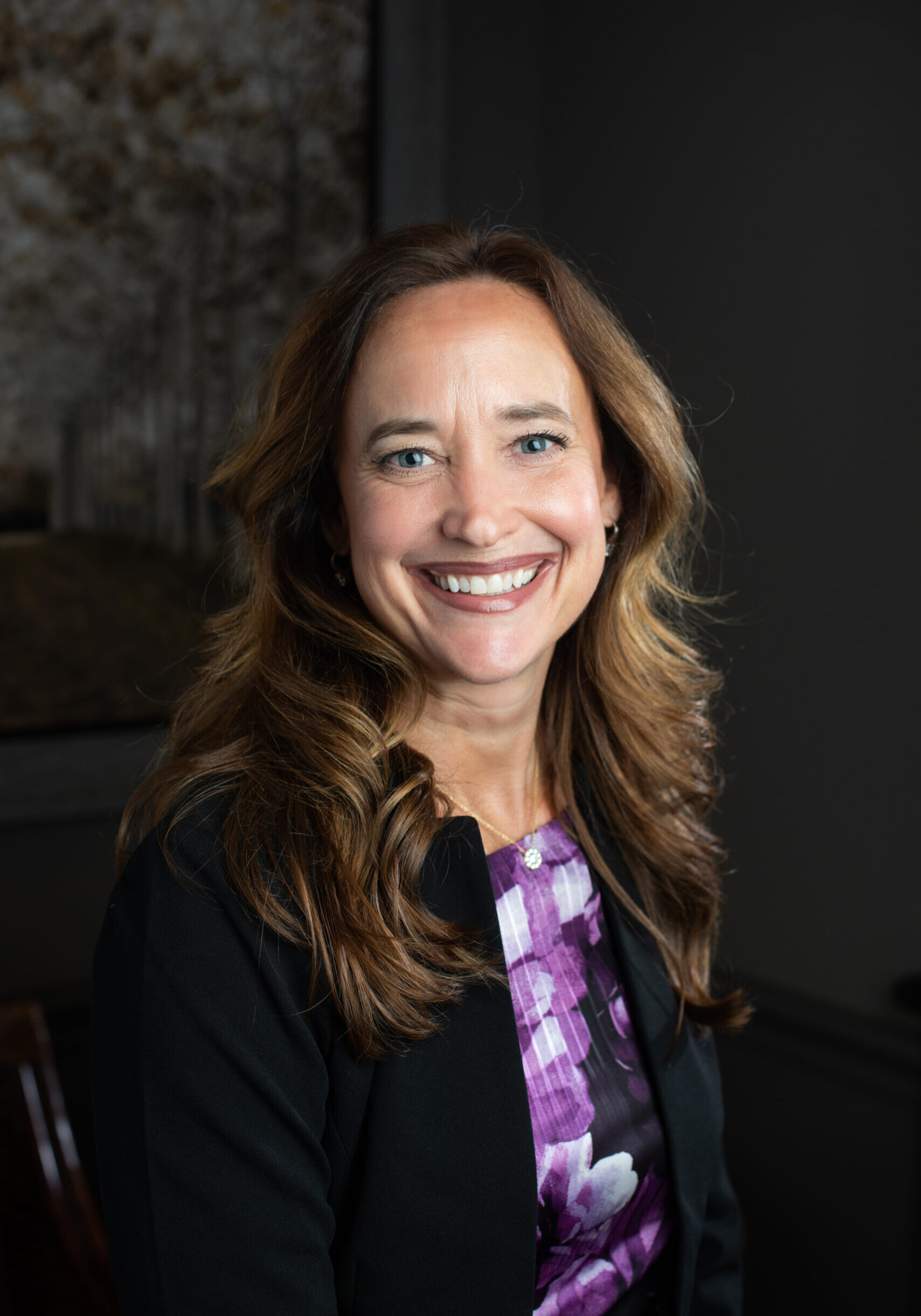 Portrait of a smiling woman with wavy brown hair wearing a purple patterned blouse and black blazer, indoors.